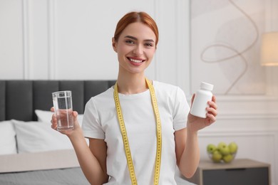 Photo of Happy young woman with bottle of pills, water and measuring tape at home. Weight loss