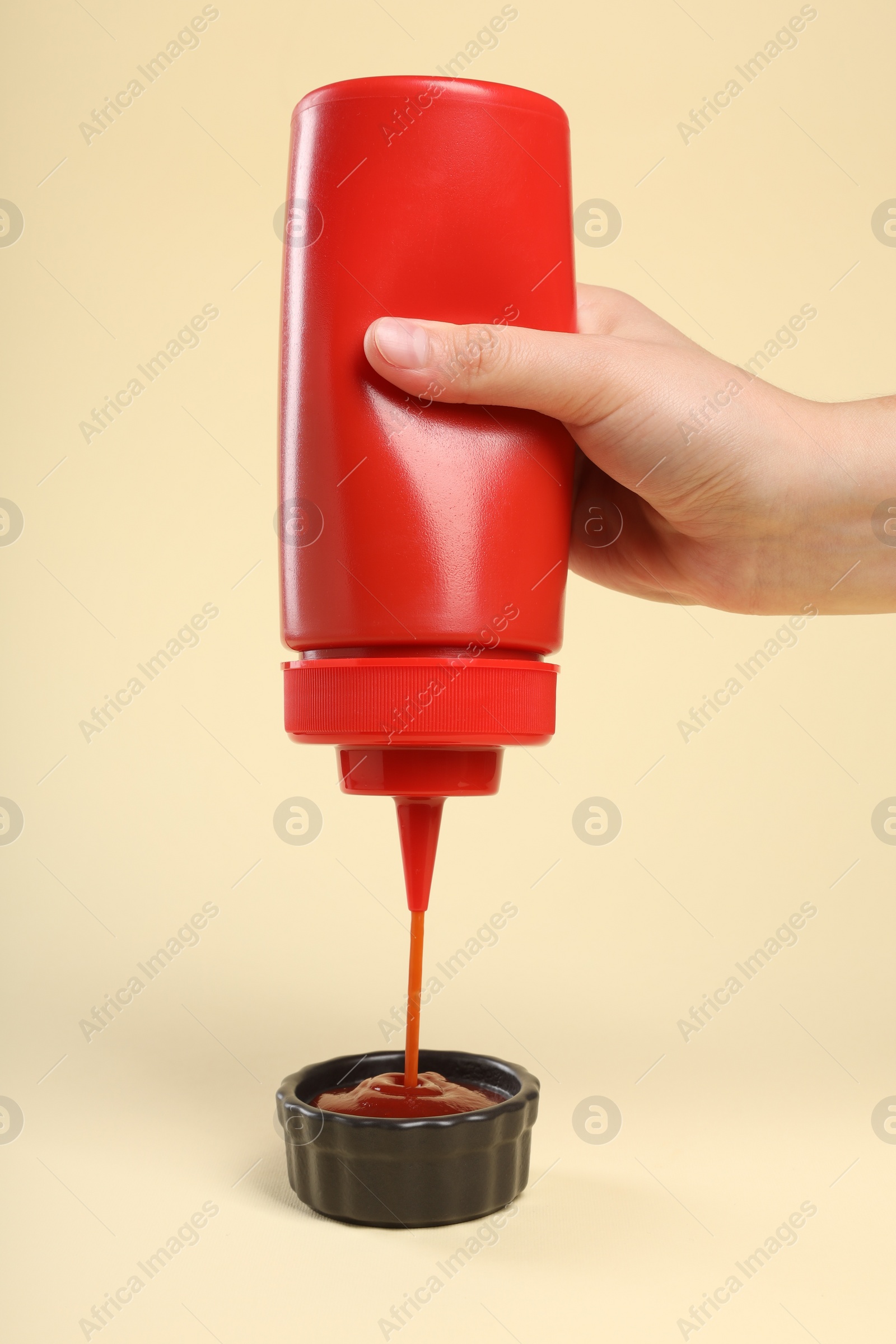 Photo of Woman squeezing tasty ketchup from bottle into bowl on beige background, closeup