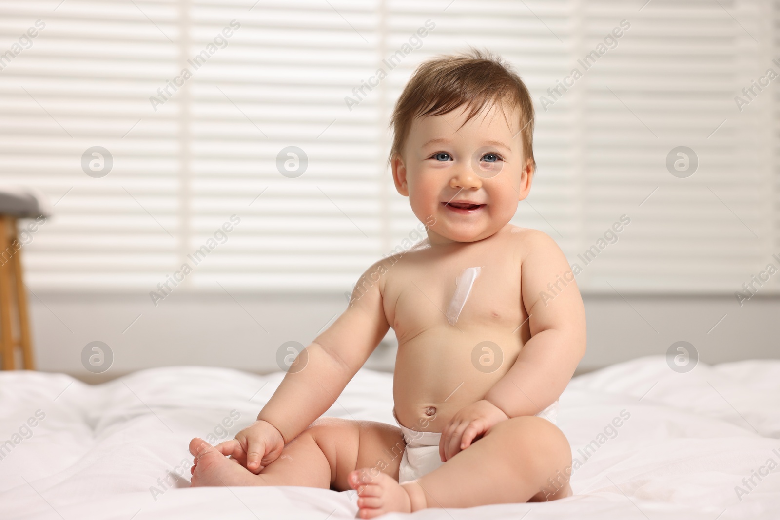 Photo of Cute little baby with moisturizing cream onto body sitting on bed at home