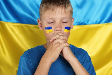 Photo of Little boy with clasped hands praying for Ukraine near national flag. No war concept