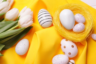 Photo of Many painted Easter eggs and tulip flowers on yellow cloth, above view