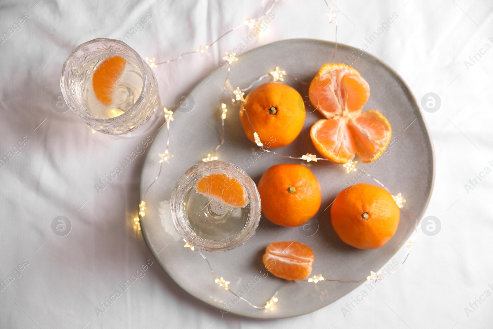 Photo of Delicious ripe tangerines, glasses of sparkling wine and fairy lights on white bedsheet, flat lay