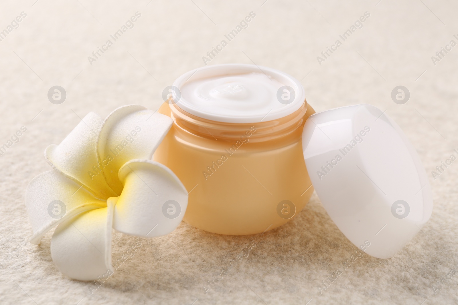 Photo of Body care. Moisturizing cream in open jar and flower on light textured table, closeup