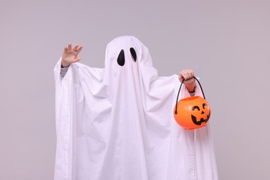 Child in white ghost costume holding pumpkin bucket on light grey background. Halloween celebration