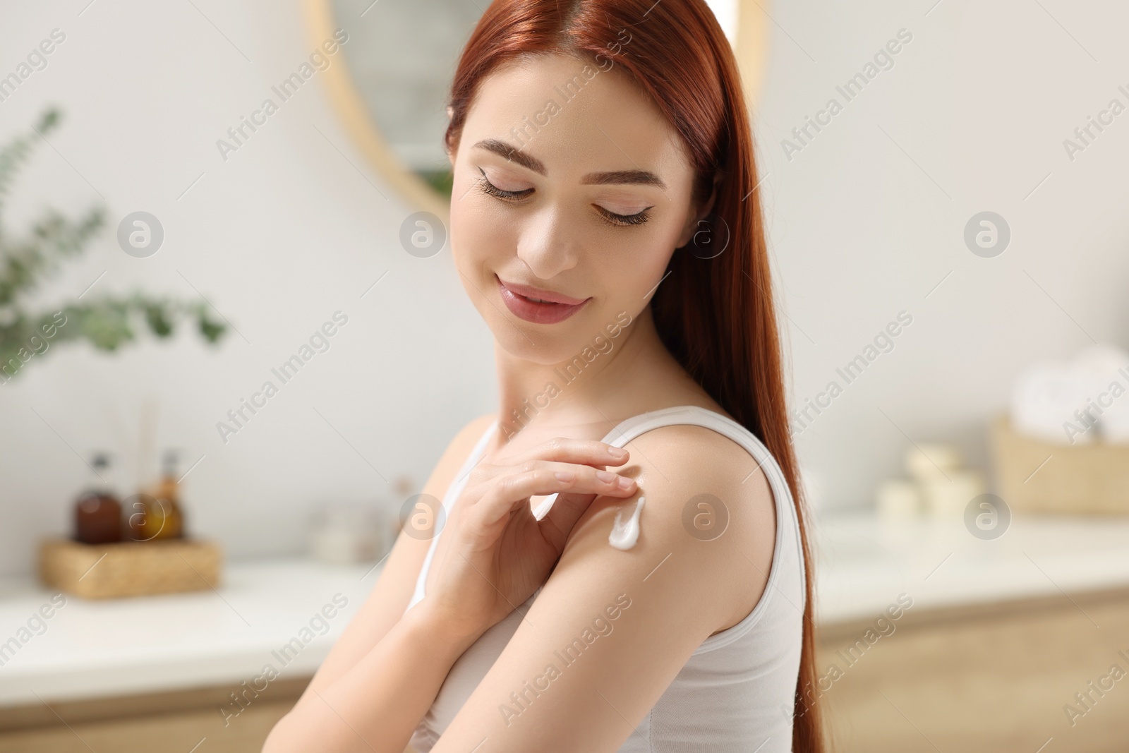 Photo of Beautiful young woman applying body cream onto shoulder in bathroom