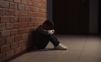 Photo of Sad little boy sitting on floor near brick wall indoors