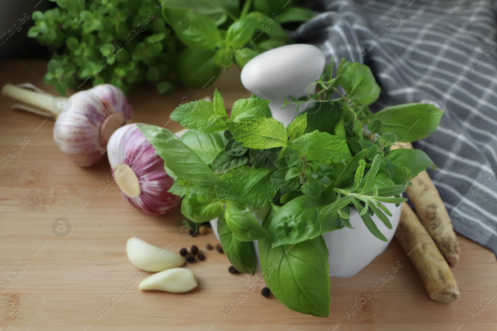 Photo of Mortar with different fresh herbs near garlic, horseradish roots and black peppercorns on wooden table