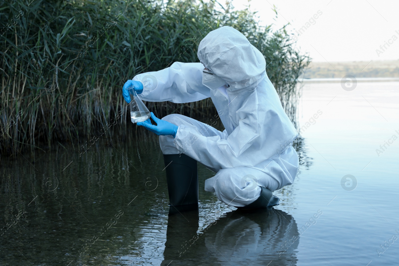 Photo of Scientist in chemical protective suit with conical flask taking sample from river for analysis