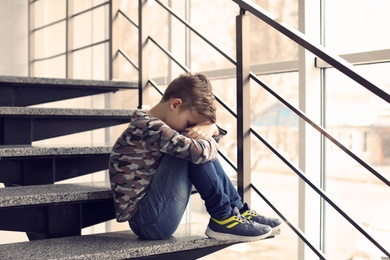 Photo of Depressed little boy sitting on stairs indoors. Time to visit child psychologist