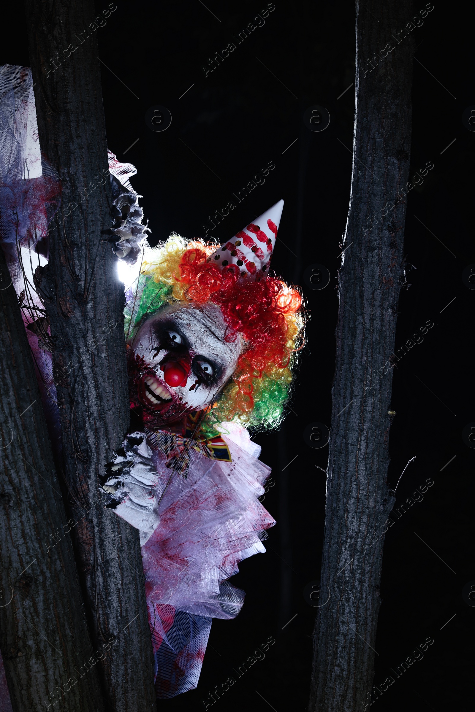 Photo of Terrifying clown hiding behind trees outdoors at night. Halloween party costume