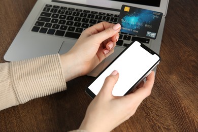 Photo of Online payment. Woman using smartphone and credit card near laptop at wooden table, closeup