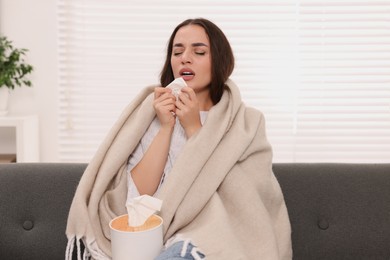 Photo of Sick woman wrapped in blanket with tissue sneezing on sofa at home. Cold symptoms
