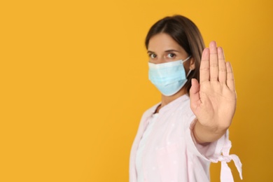 Photo of Woman in protective mask showing stop gesture on yellow background, space for text. Prevent spreading of coronavirus
