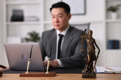 Photo of Notary working with laptop at wooden table in office, focus on statue of Lady Justice