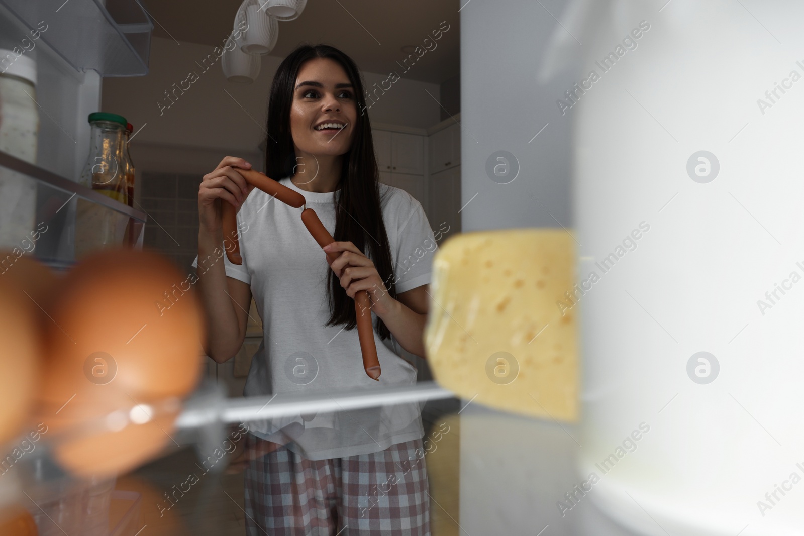 Photo of Young woman with sausages near modern refrigerator in kitchen at night, view from inside