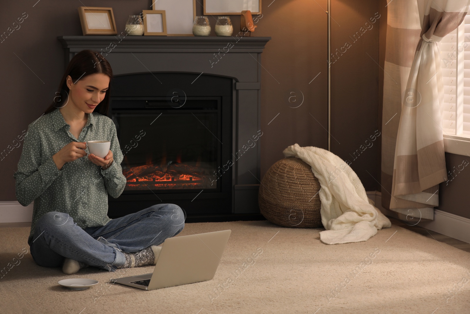 Photo of Beautiful young woman with laptop and cup of hot drink on floor near fireplace at home. Space for text