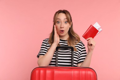 Emotional young woman with passport, ticket and suitcase on pink background