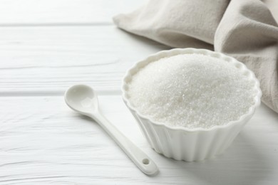Photo of Granulated sugar in bowl and spoon on white wooden table, closeup. Space for text