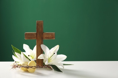 Photo of Wooden cross, crown of thorns, Easter eggs and blossom lilies on table against color background, space for text