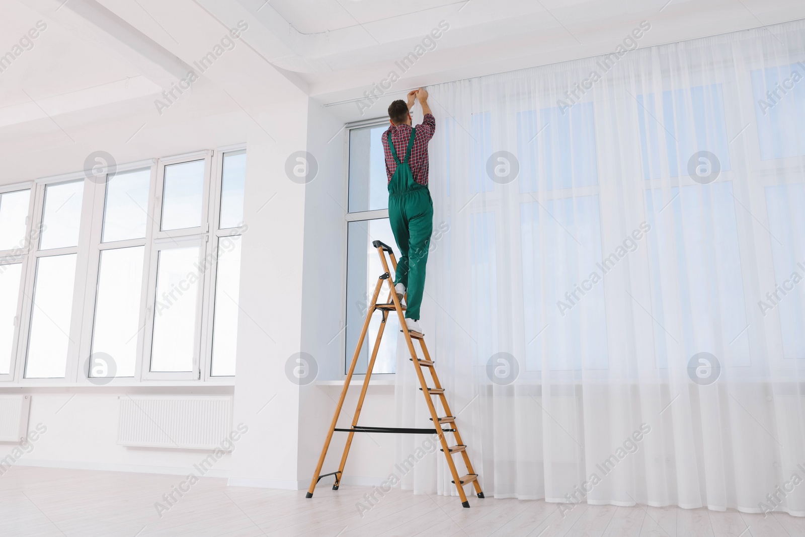 Photo of Worker in uniform hanging window curtain indoors, back view