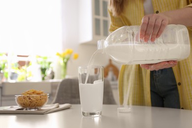 Photo of Young woman pouring milk from gallon bottle into glass at white table in kitchen, closeup