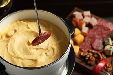 Photo of Dipping piece of sausage into fondue pot with melted cheese at table, closeup