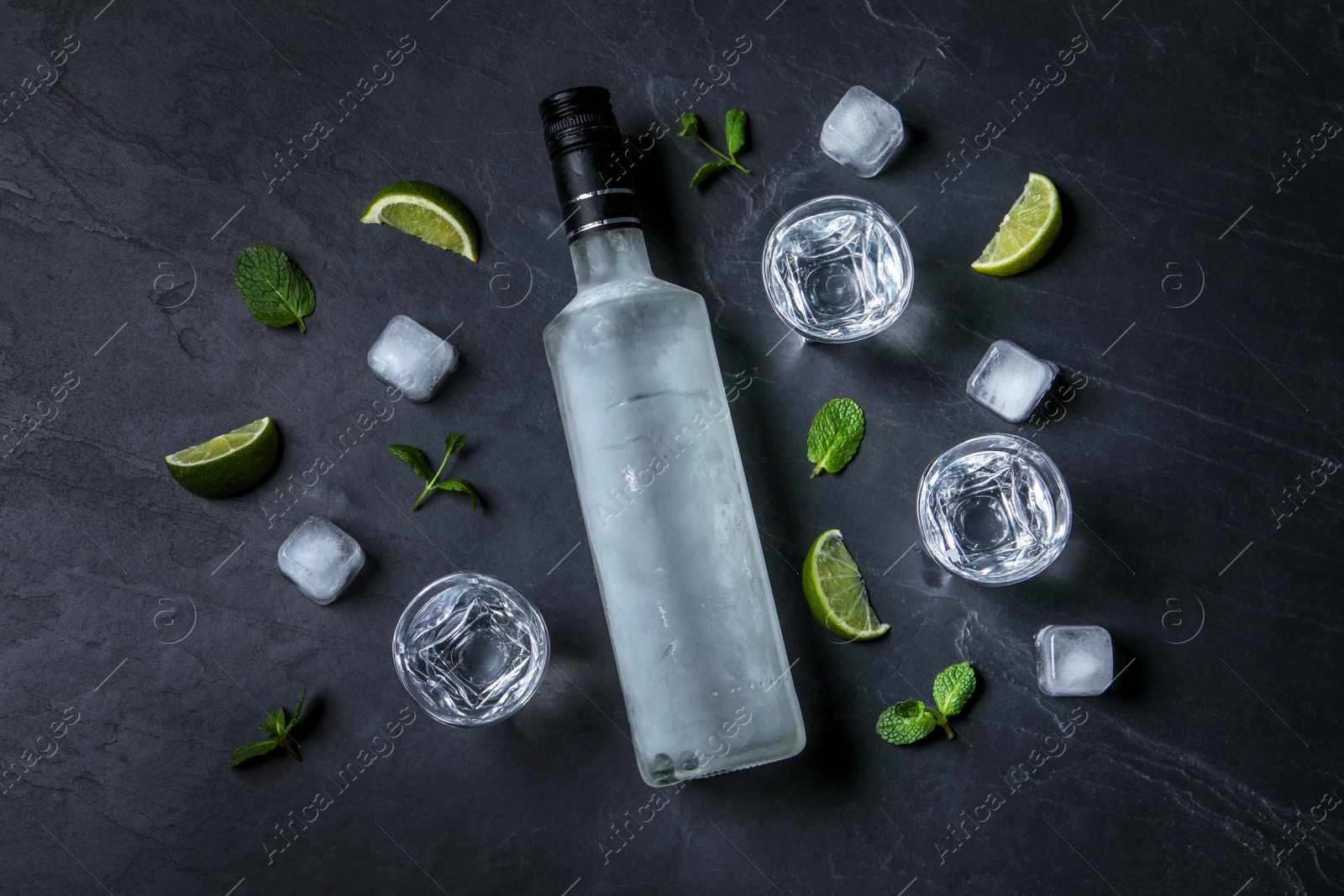 Photo of Bottle of vodka, shot glasses, lime, mint and ice on black table, flat lay
