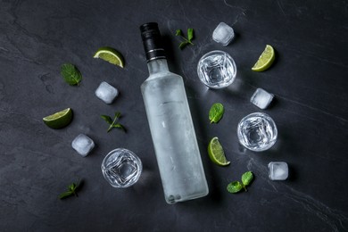 Bottle of vodka, shot glasses, lime, mint and ice on black table, flat lay