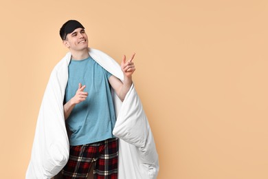 Photo of Happy man in pyjama and sleep mask wrapped in blanket on beige background, space for text