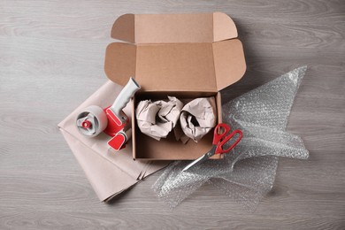 Photo of Open box with wrapped items, adhesive tape, scissors, paper and bubble wrap on wooden table, flat lay