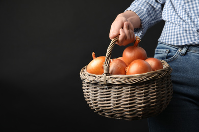 Photo of Woman holding wicker basket with raw yellow onion bulbs on black background, closeup. Space for text