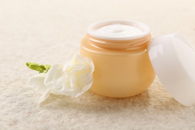 Photo of Body care. Moisturizing cream in open jar and flower on light textured table, closeup