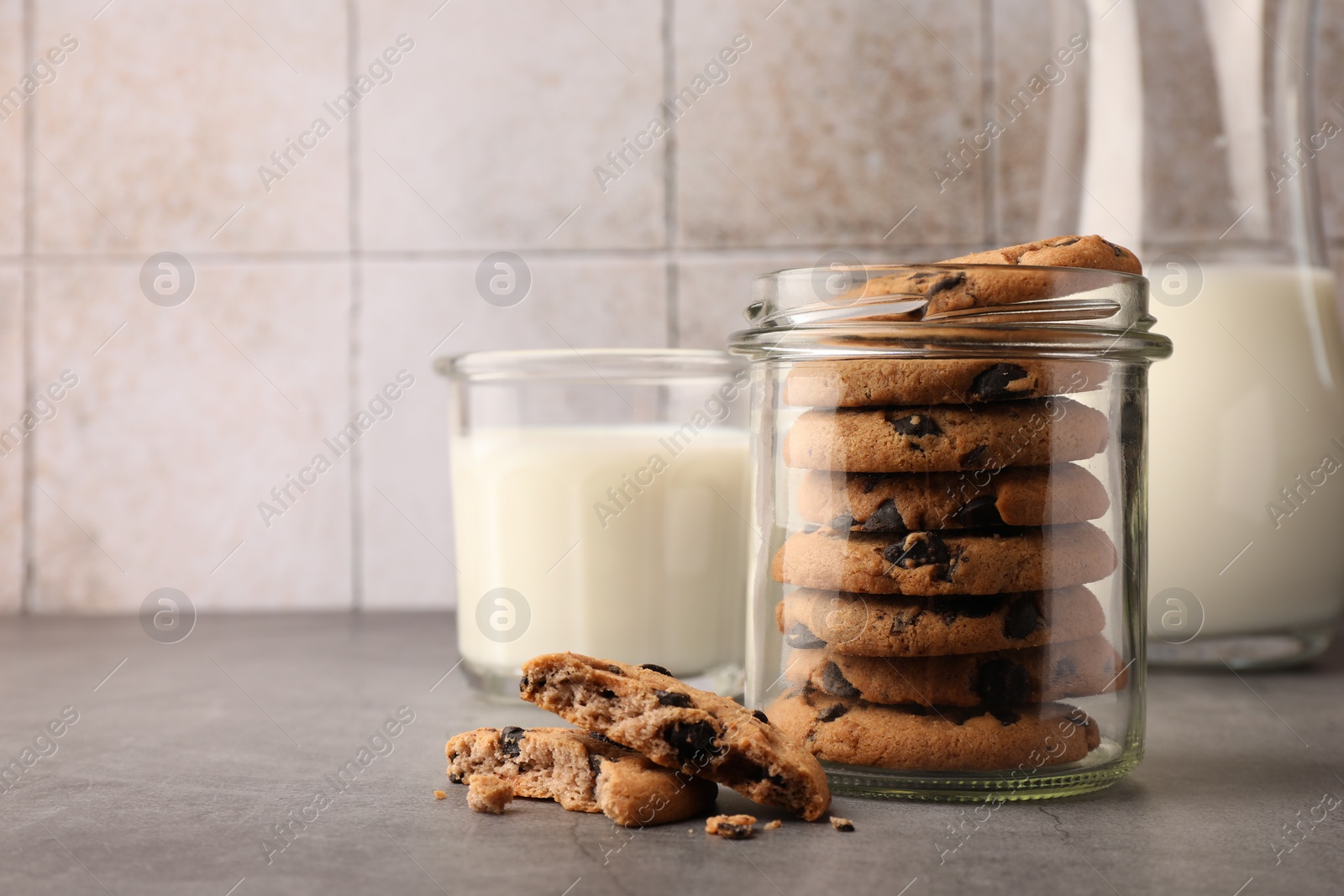 Photo of Glass jar with delicious chocolate chip cookies and milk on grey table. Space for text