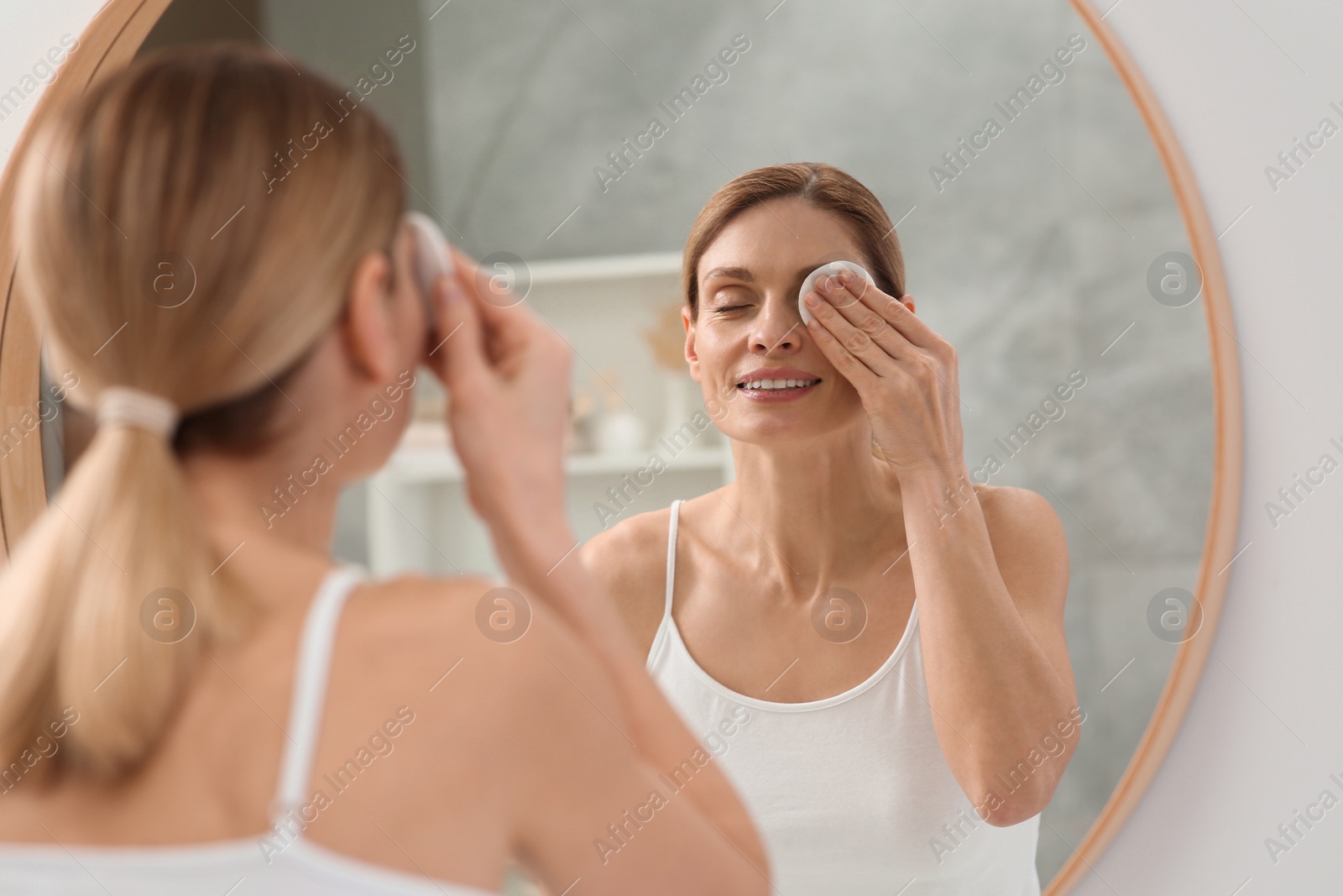 Photo of Beautiful woman removing makeup with cotton pad near mirror indoors