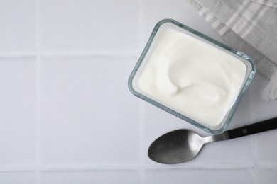 Delicious natural yogurt in bowl and spoon on white tiled table, top view. Space for text