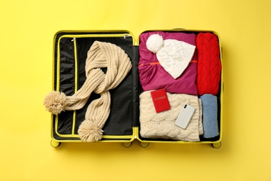 Open suitcase with warm clothes, passport and smartphone on color background, top view