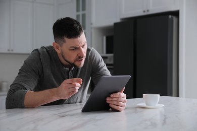 Photo of Emotional man with tablet at table in kitchen. Online hate concept