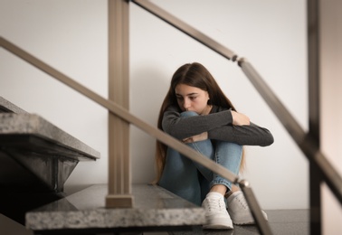 Photo of Upset teenage girl sitting on stairs indoors. Space for text