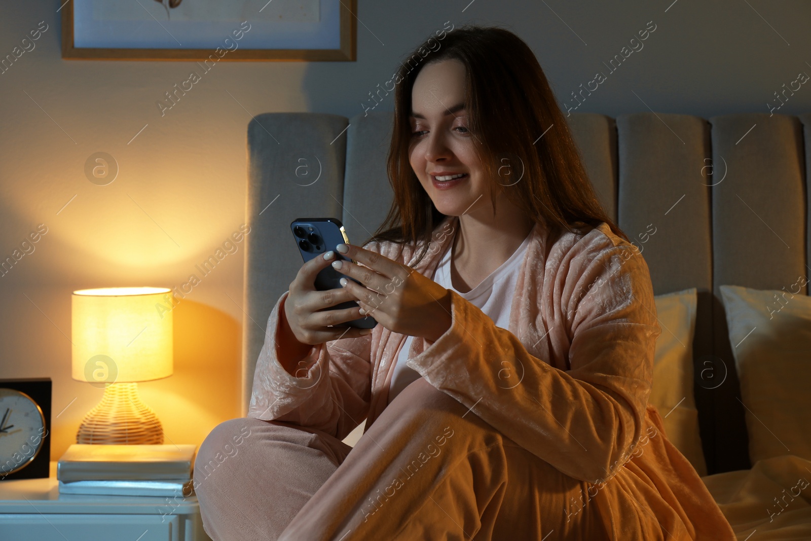 Photo of Happy woman with smartphone in bedroom at night. Internet addiction