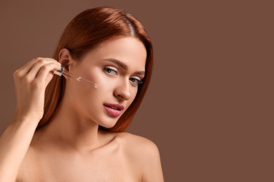 Beautiful young woman applying cosmetic serum onto her face on brown background, space for text