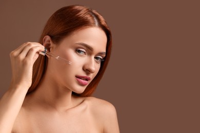 Photo of Beautiful young woman applying cosmetic serum onto her face on brown background, space for text