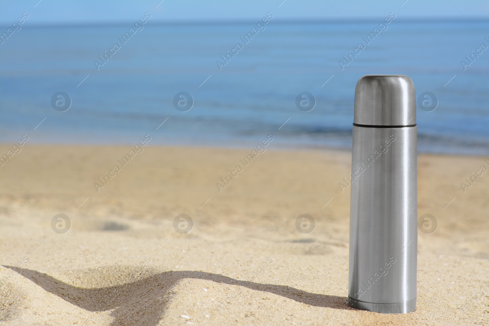 Photo of Metallic thermos with hot drink on sandy beach near sea, space for text