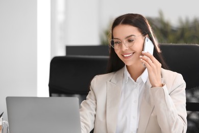 Happy woman using modern laptop while talking on smartphone in office