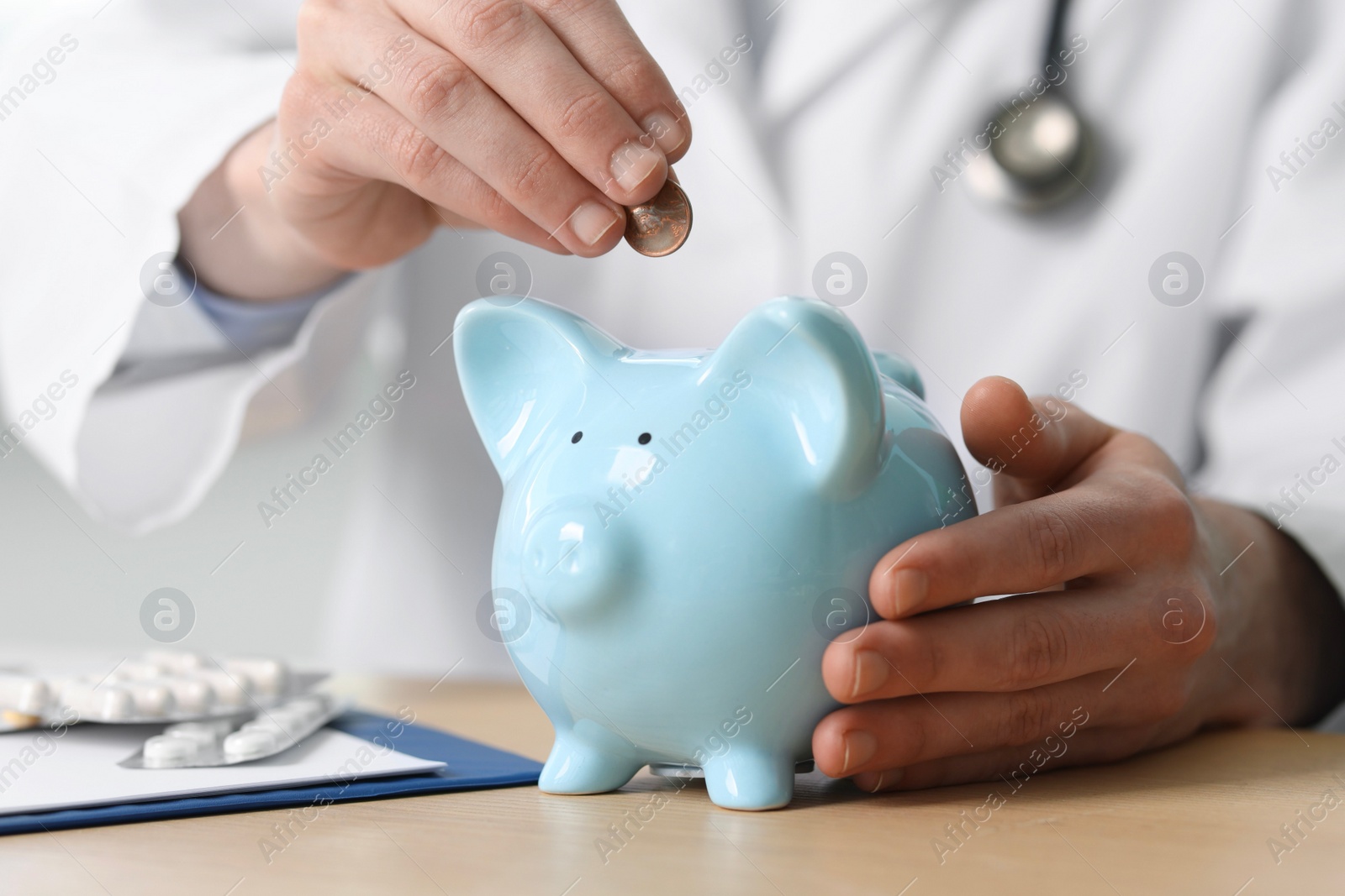 Photo of Doctor putting coin into piggy bank at wooden table, closeup