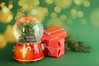 Image of Beautiful Christmas snow globe, gift boxes and fir tree branch on green background, bokeh effect. Space for text