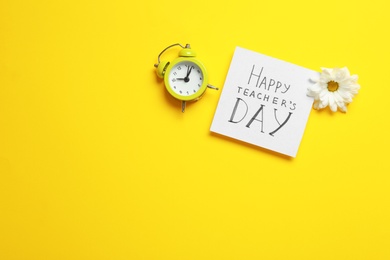 Flat lay composition of card with inscription HAPPY TEACHER'S DAY, flower and alarm clock on yellow background, space for text