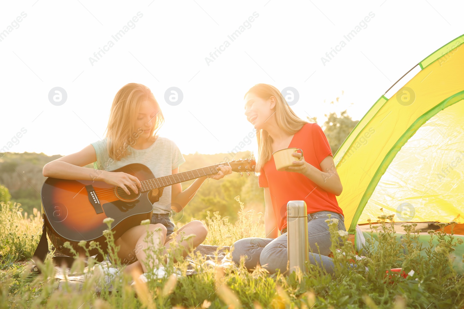 Photo of Young women resting with hot drink and guitar near camping tent in wilderness