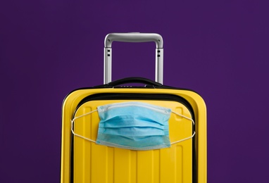 Stylish yellow suitcase with protective mask on purple background. Travelling during coronavirus pandemic