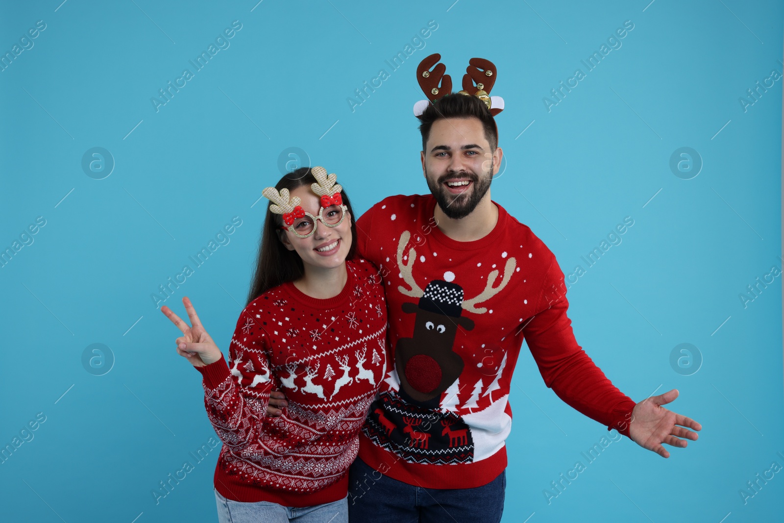 Photo of Happy young couple in Christmas sweaters, reindeer headband and funny glasses on light blue background