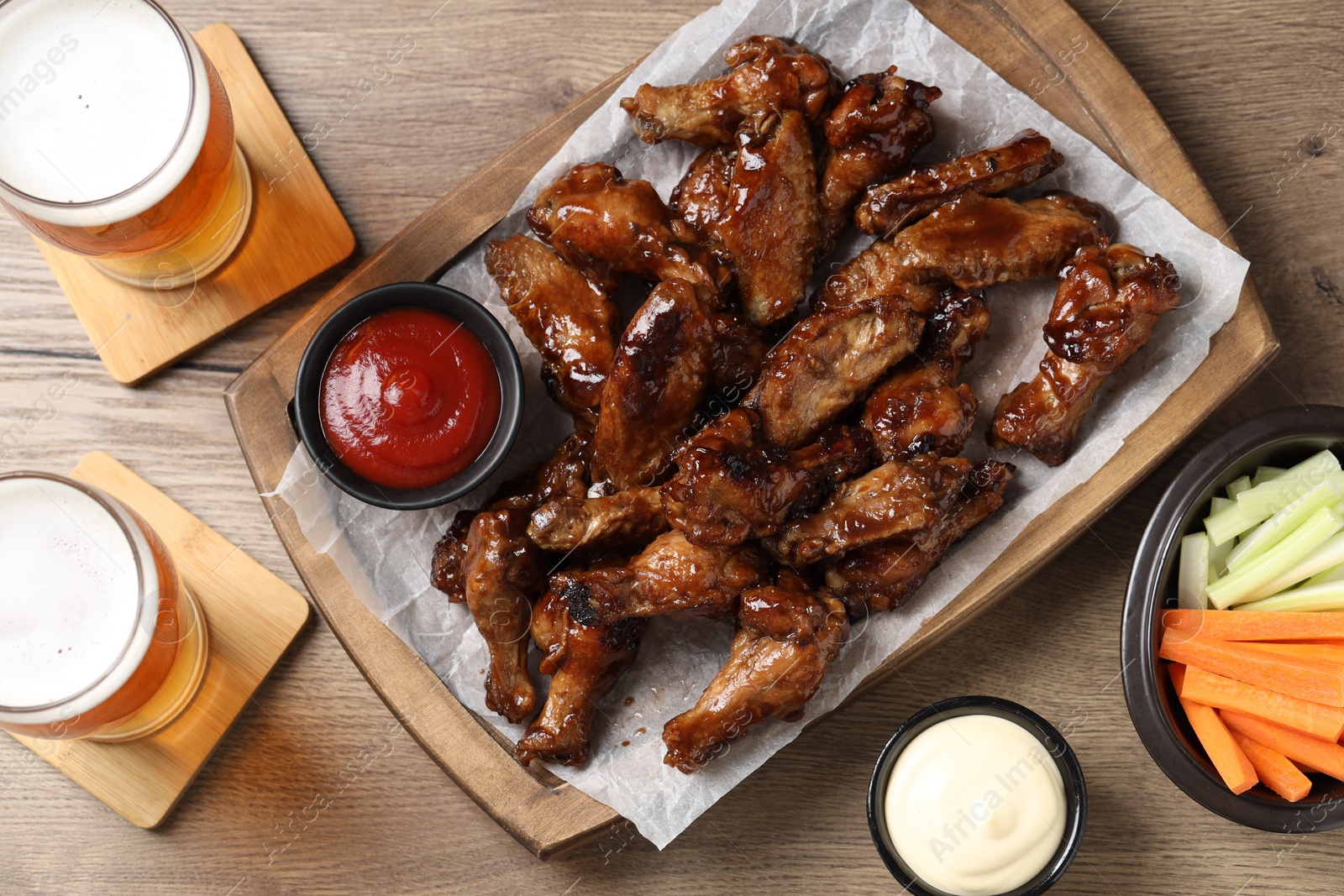 Photo of Tasty chicken wings, glasses of beer, sauces and vegetable sticks on wooden table, flat lay. Delicious snacks